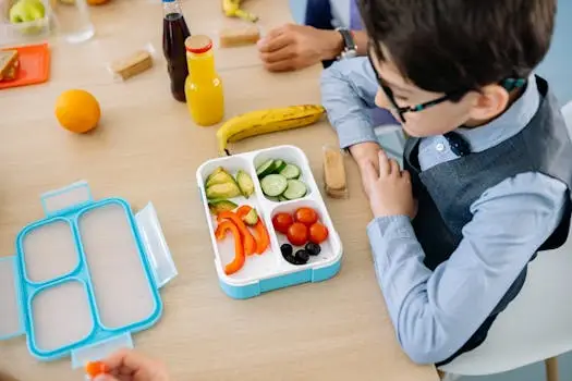 A young boy sits at a wooden table in school, enjoying a healthy lunch from a colorful lunchbox.